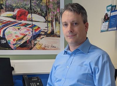   Gavan Duffy, Advanced Nurse Practitioner in Movement Disorders. Gavan is seated in an office. He is waering a blue shirt. A large colourful painting is visible behind him. 