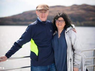 A man and a woman standing in front of a railing outside with the sea behind them.