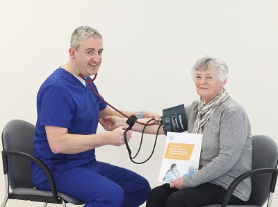 A doctor wearing blue scrubs and a stetascope is seated with a female patient. She is holding a Chronic Disease Management leaflet while he is taking her blood pressure. 