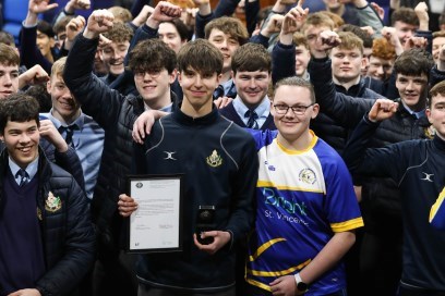 Noah Winders is holding his bravery award and standing beside his friend Charlie Rutter. Charlie has his arm around Noah's shoulders.  Behind them are a large group of other boys in their school uniforms. 