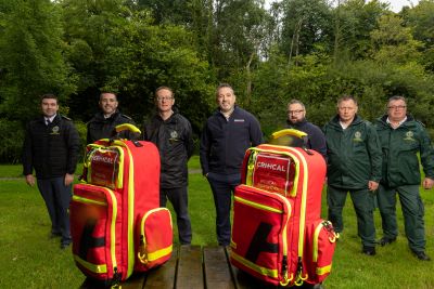 7 Members of the National Ambulance Service and charity CRITICAL standing outside in a leafy area. In front of them are 2 large  red CRITICAL medical bags 