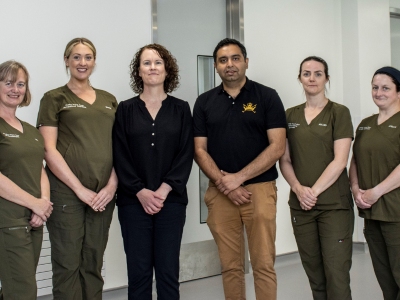 Five women and one man standing indoors, four of the women wear green medical scrubs.