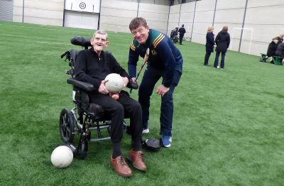 A smiling service user is seated in a wheelchair holding a football. Aa coach holding a hurl is leaning in beside smiling.
