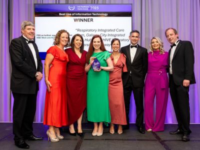 Eight people in formal clothes standing on a stage in front of a TV screen. A woman in the centre in a green dress is holding a plaque.