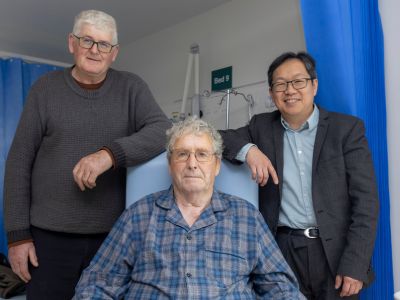 Three men in a clinical setting, one is sitting down and the other two stand on either side of him.