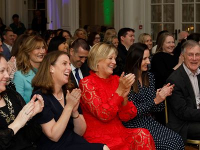 A crowd of people wearing formal clothes seated at an indoor event. They are all looking ahead, smiling and applauding.