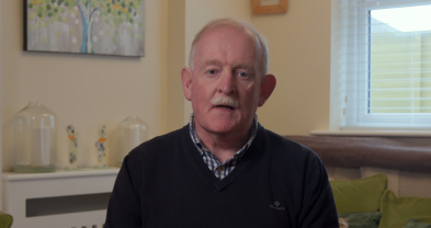 Martin Gillick is seated on a couch in a sitting room. He is looking straight ahead. Behind him is a window and there and painting on the wall and vases on the mantelpiece.