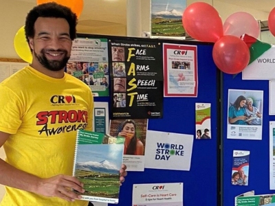 A man wearing a stroke awareness t-shirt, holding a booklet and standing beside a stand covered in stroke awareness graphics.