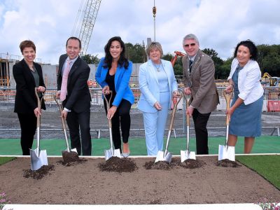 Six people in formal clothes holding shovels over a plot of dirt outdoors.