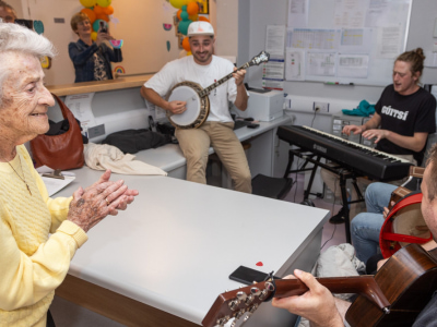 A group of musicians playing musical instruments in a room while an older woman watches and claps her hands.