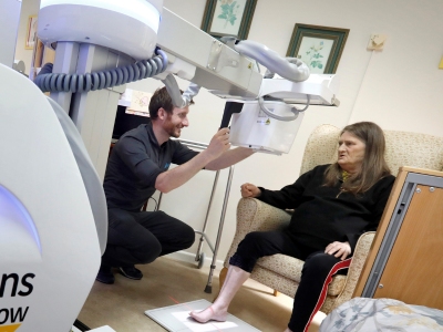 An older woman sits on an armchair while a younger man interacts with a large mobile x-ray machine setup in front of her.
