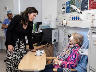 A woman standing and speaking to an older patient in a clinical setting. The patient has a white patch over her eye and there is medical equipment on either side of her. Behind them is a healthcare worker in blue medical clothes.
