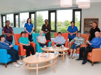 Six older people sitting in armchairs indoors, with five healthcare workers standing behind them.