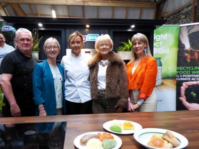 A woman in a chef's uniform stands with four people in regular clothes behind plates of food on a countertop.