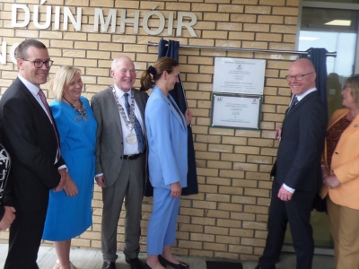 Minister Stephen Donnelly standing beside a plaque on the wall of a healthcare setting with two other men and three women wearing formal clothes.
