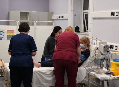 3 healthcare workers are standing around a female patients' hospital bed. 2 of the healthcare workers are providing care to the patient