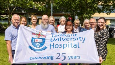 A large group of staff and management from Tallaght University Hospital holding a large white flag with the words 'Tallaght University Hospital celebrating 25 years'