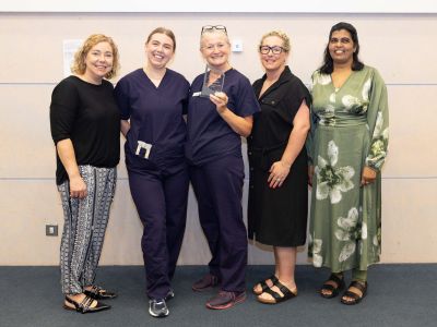 Five women standing in a row and smiling. Two in the centre are wearing medical clothes and one is holding a small plaque.