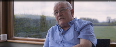 Sean Crombie is seated. He is wearing a blue shirt and glasses. There are windows behind him with views out to fields. 