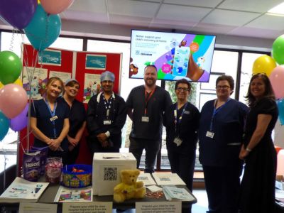Seven hospital staff members stand behind a table with various informational materials on it. There are baloons beside them and more informational material on a board behind them.
