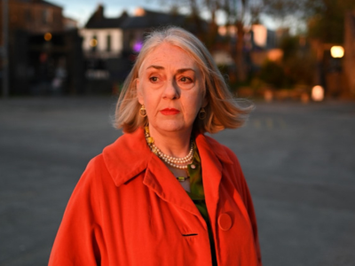 A woman with grey hair and a red coat stands outside with some houses and trees in the background.