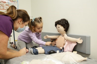 A doll is on a hospital bed. A nurse is leaning over the bed while a young girl of about 4 checks the doll's chest. 