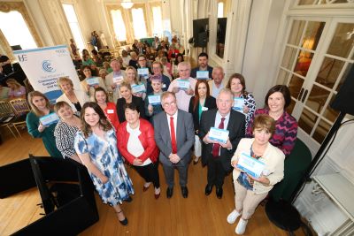 Very large group of people in  a formal function room. Some members of the group are holding small blue cards that read, Community Cancer Support Centres and Services. A pull-up banner is also visible in the background.