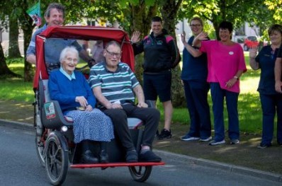 Male Trishaw pilot from Killarney Cycling Club taking  2 male resiidents, 1 male and i female on a cycle in the trishaw. A small crowd waves as they pass.    