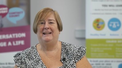 A smiling Carmel Dunne looks straight ahead. In the background are health promotion posters.