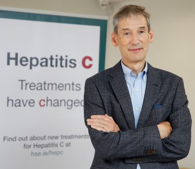 Professor Aiden McCormick, HSE Clinical Lead for the Hepatitis C Programme standing with folded arms in front of a sign that reads' Hepatitis C Treatments have changed'