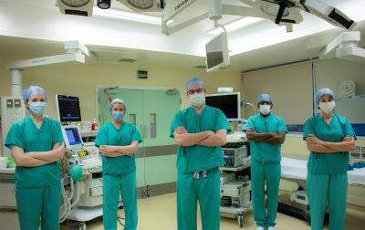 A group of clinicians in green scrubs and masks stand with arms folded in a theatre setting