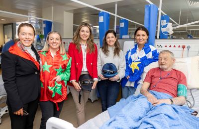 A group of 5 young female jockeys are in a row and smiling at the hospital bedside of a male patient 