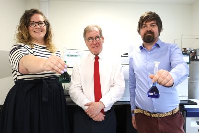 3 people standing in a row lab setting. A man in a red tie stands in the middle while a woman on one side and a man on the other are holding out test tubes