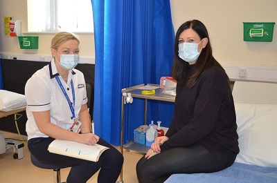 Patient sitting on hospital bed looking towards the camera. Beside her a nurse is seated. She is also looking at the camera.