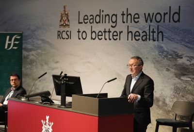 Dr Colm Henry, HSE Chief Clinical Officer standing at a podium. Behind him is the RCSI logo and the words 'Leading the world to better health'.