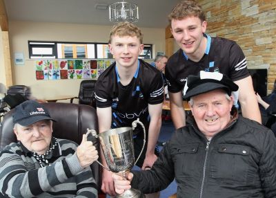 2 members of the Sligo GAA under 20s team stand holding the JJ Fahy Cup. Seated are 2 male residents of residents of St John’s Hospital , Sligo who are also holding the cup