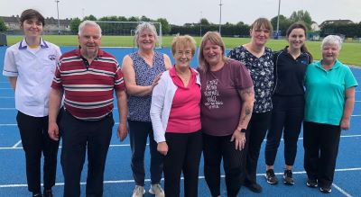 Large group of people standing in a line on an athletics track. There are a couple of healthcare workers with the group. 