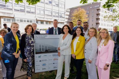 Large group of people standing around a sign depicting an image of a building. There are tall buildings in the background. 