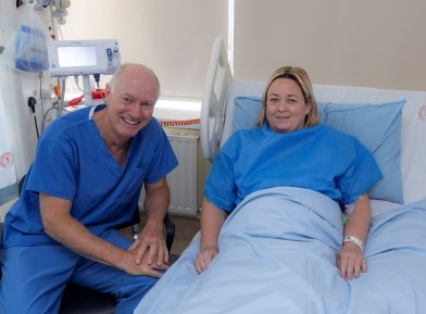 A doctor in blue scrubs is seated beside a hospital bed with a female patient in it. Both are smiling.