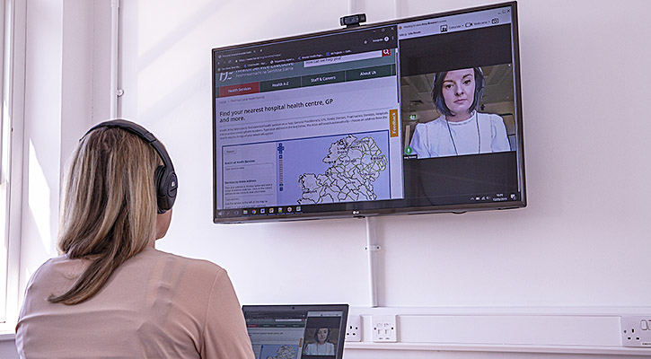 Photo of Ulla and Amy demonstrating how they conduct remote online usability testing with users. Ulla is seated in the office in front of her laptop with headphones on and Amy's face is appearing on a large monitor on the wall.
