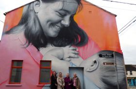 Four women standing outdoors in front of a large mural of a woman breastfeeding a baby.