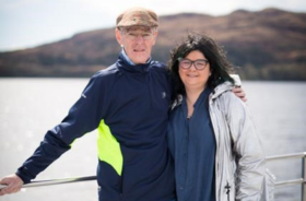 A man and a woman standing in front of a railing outside, with the sea behind them.