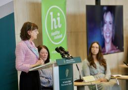 Bernie Rice speaking a podium. The Healthy Ireland logo is in the background. A powerspoint presenation with a photo of Bernie's daughter Sharon is also visible in the background.
