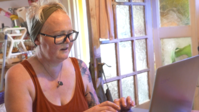 Celine Garvey seiiting in her kitchen. She is wearing glasses and working on a laptop.