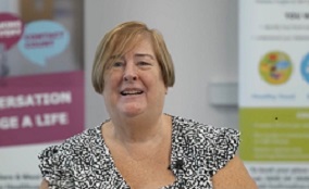 A smiling Carmel Dunne looks straight ahead. In the background are health promotion posters.