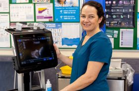 A woman in medical clothes stands beside an ultrasound machine in a clinical setting.
