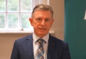 Dr Patrick Glackin is wearing a suit and tie and is standing looking straight ahead. 