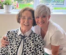 Smiling patient Kathleen O Sullivan seated in her home in Galway with GUH Frailty Team’s Therapy Assistant, Patricia Duffy who has her arm around Kathleen's shoulder.