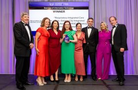 Eight people in formal clothes standing on a stage in front of a TV screen. A woman in the centre in a green dress is holding a plaque.