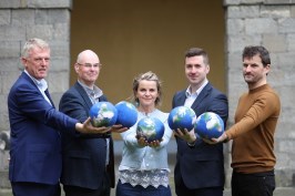A group of 5 people are standing in a semi circle a courtyard setting. They are each holding a small globe in an outstretched hand